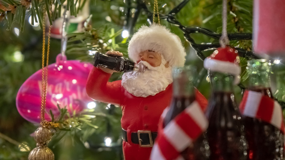 Christmas ornament of Santa Clause drinking a coke with his eyes shut. Ornament is hanging on a tree.