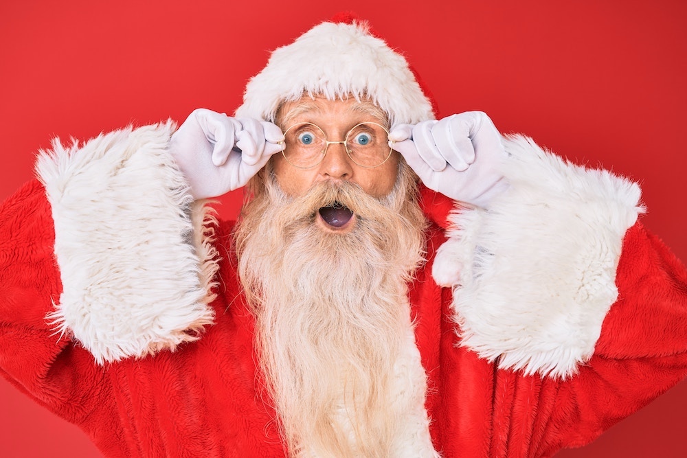 Santa Claus with hands on eyeglasses, mouth open wide, looking surprised.