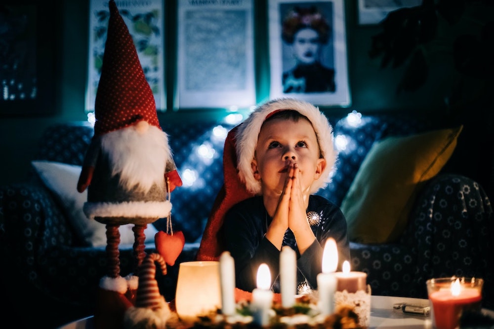 Little boy with Santa hat sitting in living room at night, hands in prayer with santa gnome standing next to him