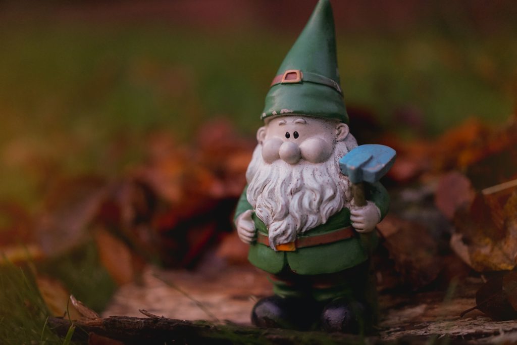 Old garden gnome in green santa suit standing in garden with garden tool
