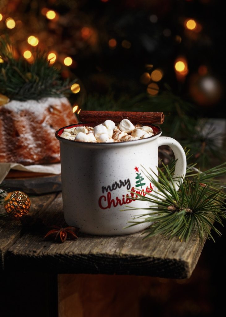 Hot cocoa in white mug with mini marshmallows on a table with Christmas branch.