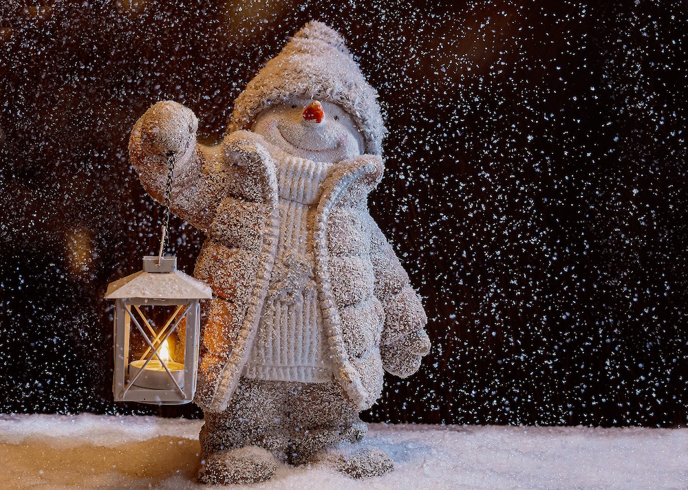 Resin snowman dressed in sweater, beige jacket, hat, mittons, pants, holder a lantern in front of black background with snow. 