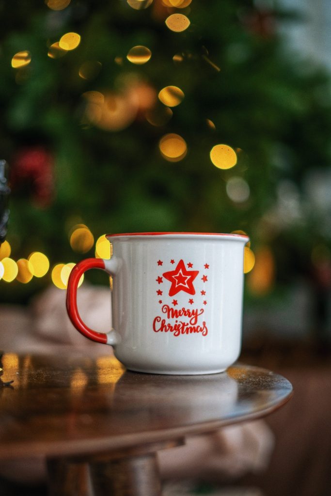 White mug with words Merry Christmas in red sitting on a table infrom of lights.