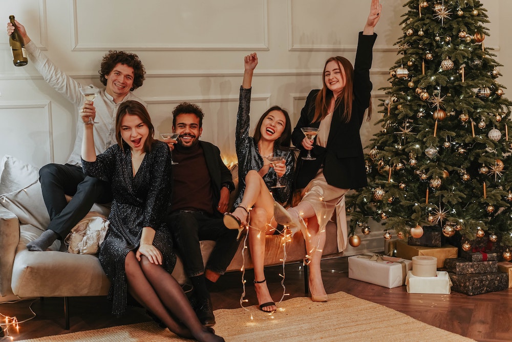 Two young men and 3 young women sitting on a couch raising their glasses next to a Christmas tree.