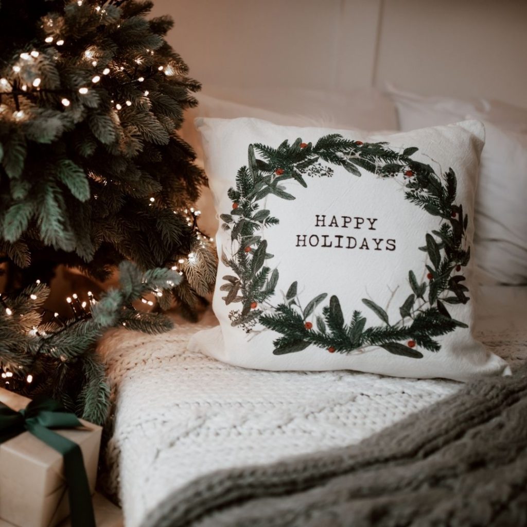 Happy holidays lettering on white pillow next to Christmas tree to show how preparing for holiday guests looks like.