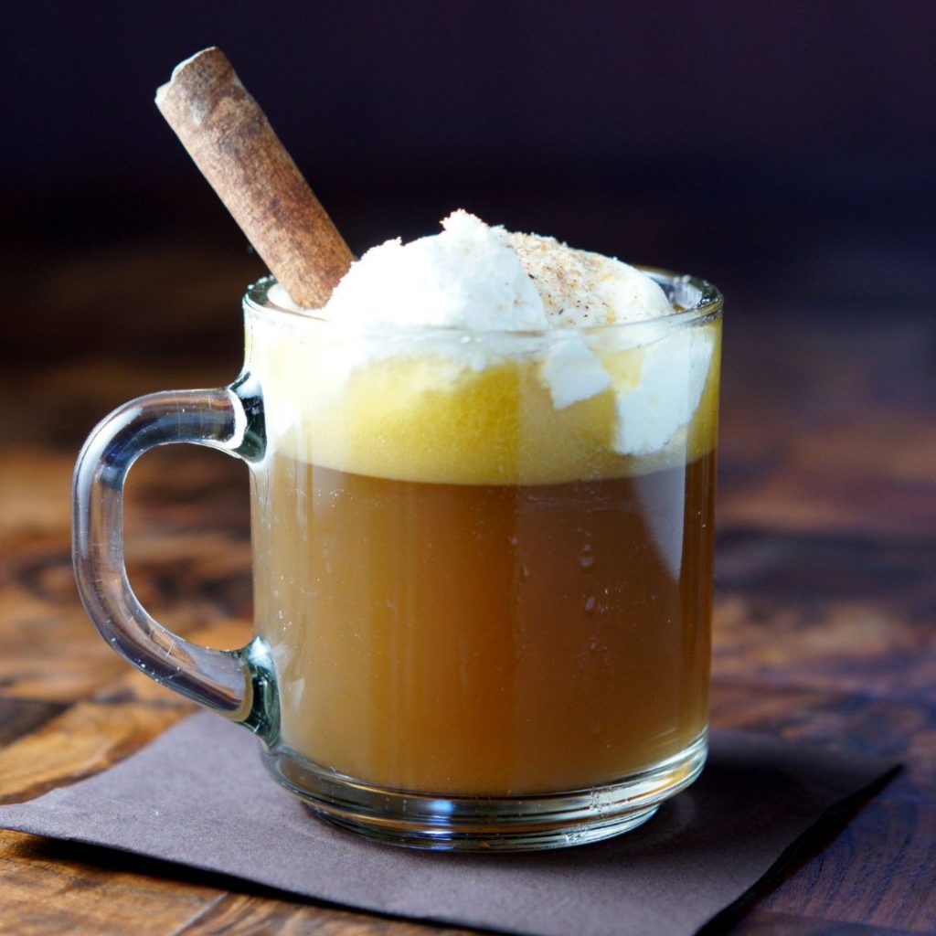 A cup of hot buttered rum in a clear mug with whipped cream and a cinnamon stick on top