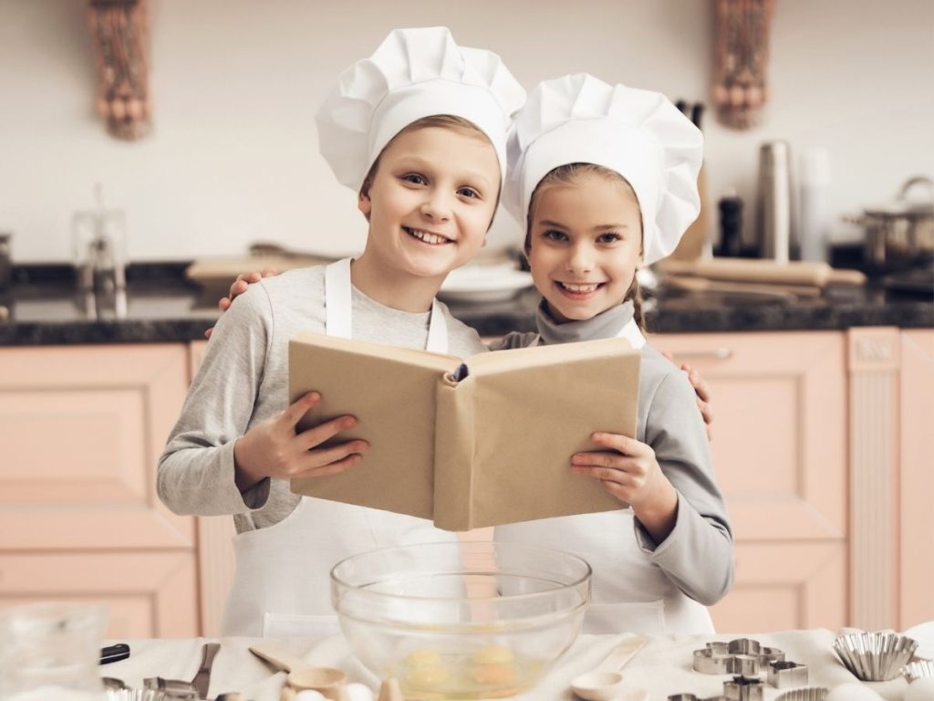 Young children with bakers hats and aprons reading family holiday cookbook together.