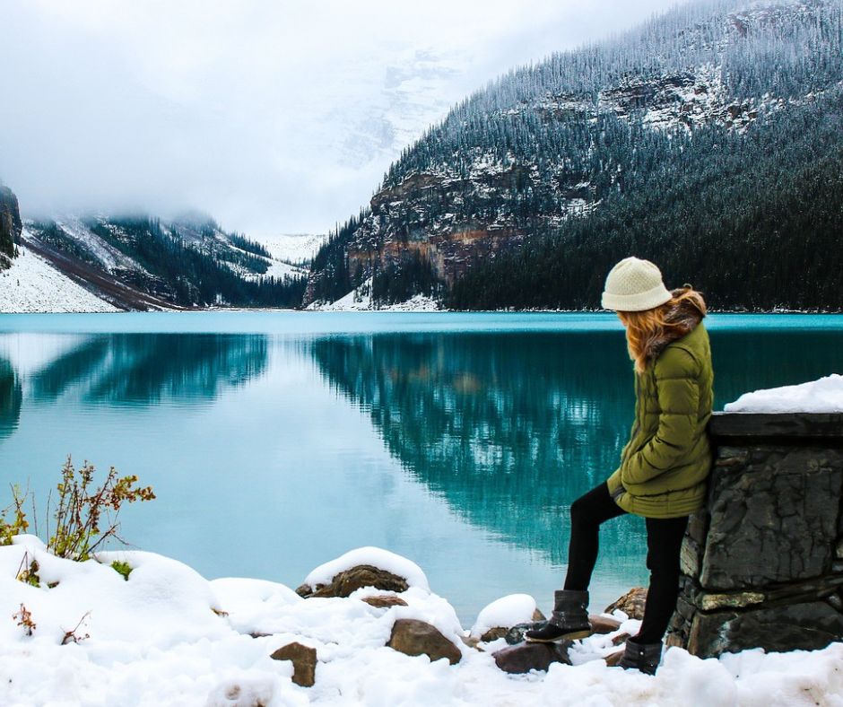 Woman standing outdoors in winter at lake with mountains in background.