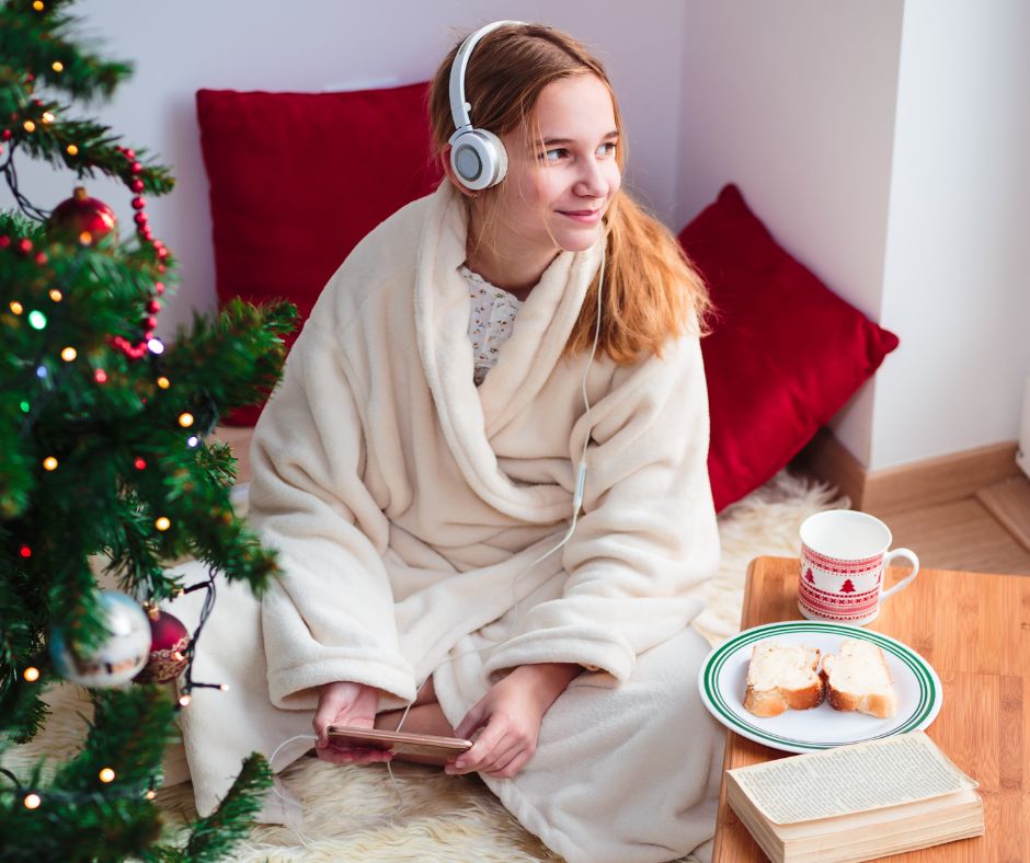 Young girl in white robe sitting on the floor in front of Christmas tree with headphones listening to Christmas Morning Christmas Podcast.